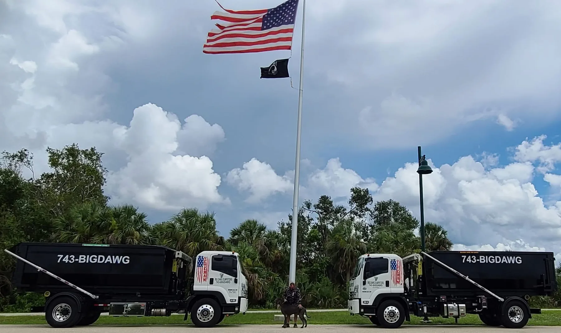Two Big Dawg roll-off trucks, Mattis the Great Dane, and a team member beneath United States and POW/MIA flags at a Southwest Florida veterans memorial.